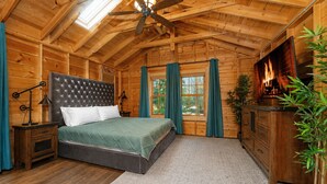 A log cabin bedroom with a bed, a dresser, a nightstand, a ceiling fan, a fireplace, and a window with curtains. - Dollybrook 1: Dreamy Den | Hot Tub & Pool (Fennville)