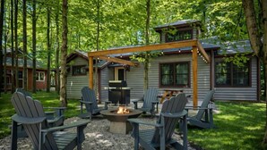 A cabin with a fire pit surrounded by Adirondack chairs, a pergola, and a deck.