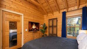 Quarto de cabana de tronco com cama, lareira, janela com cortinas e porta para outro cômodo.