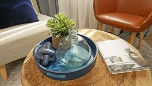 Une table ronde en bois sur laquelle repose un plateau bleu, un flacon en verre, une sculpture en verre et une plante en pot. Un magazine intitulé « COASTAL » est posé sur la table.