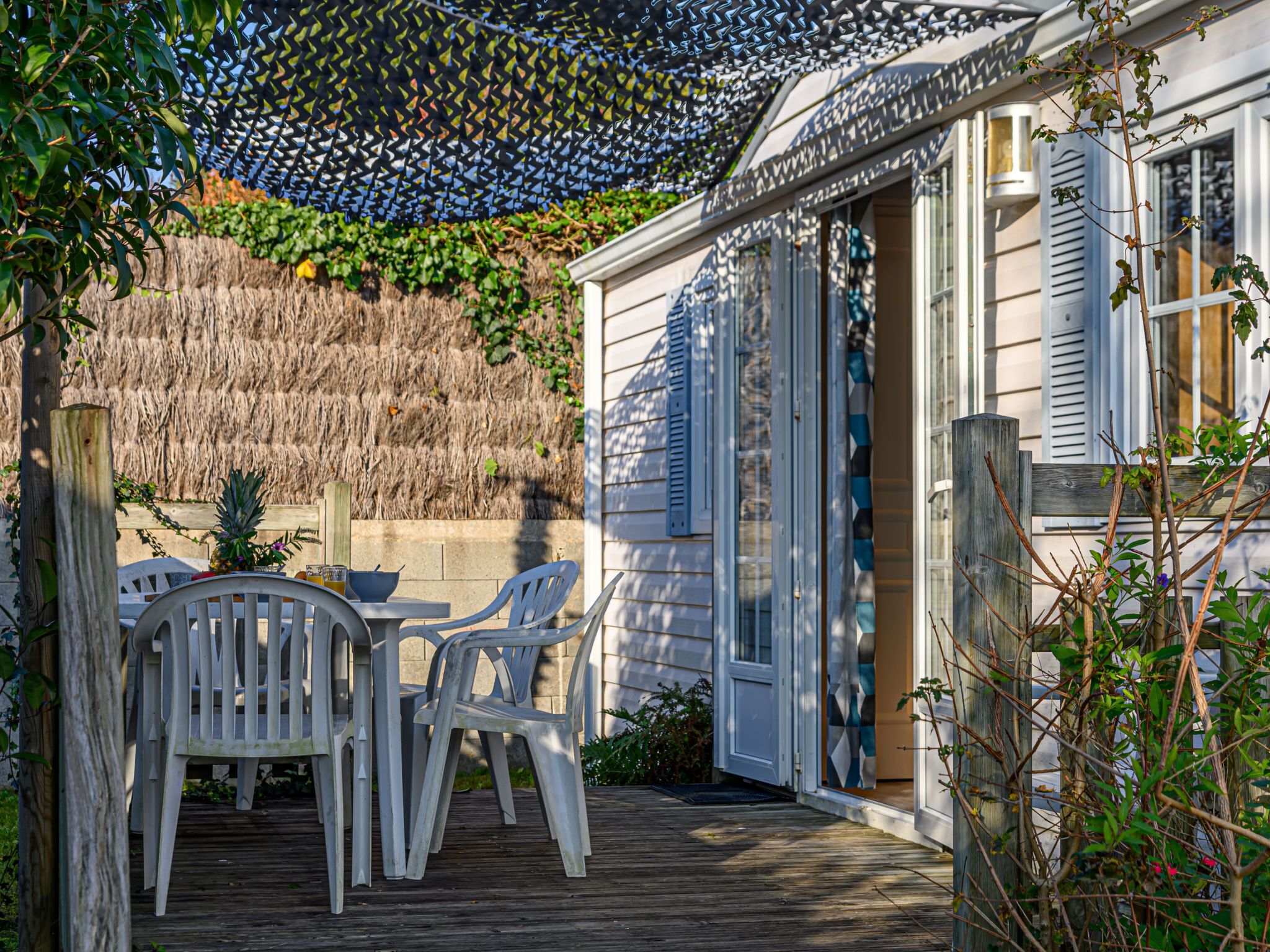 A wooden deck with a table and chairs, a thatched wall, and a house with blue shutters.