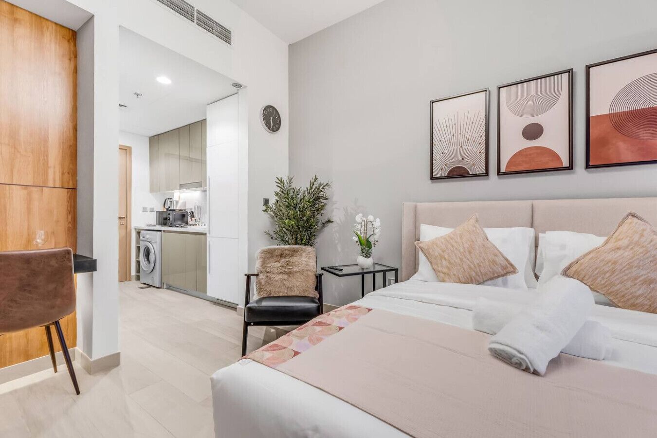 A modern hotel room with a bed, a chair, a small table, and a view into the kitchen area.