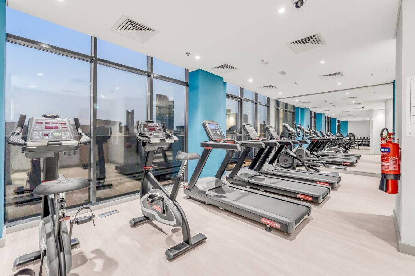 A gym with multiple treadmills and exercise bikes, large windows, and a fire extinguisher.