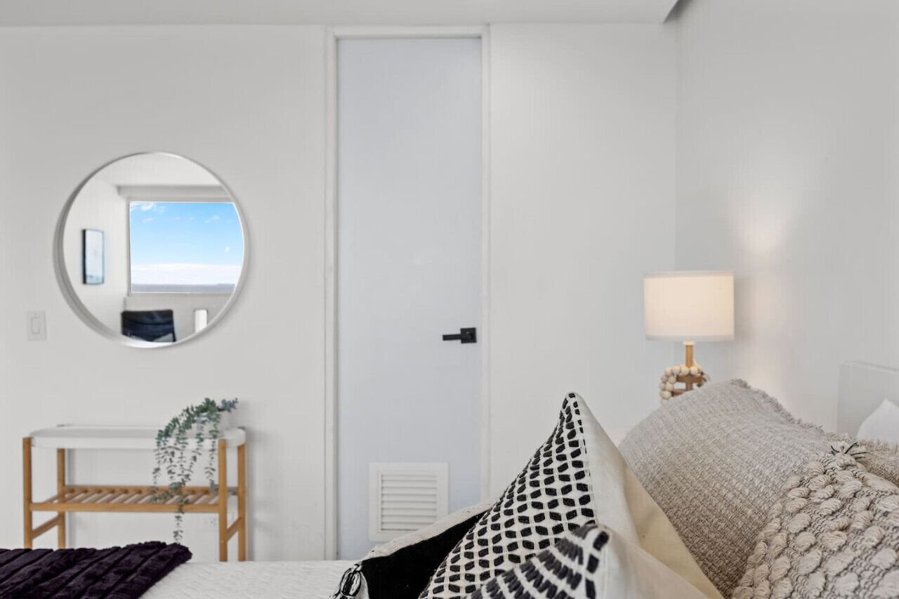 A bedroom with a bed, a bedside lamp, a round mirror, a small table with a plant, and a door with a view of the sky.
