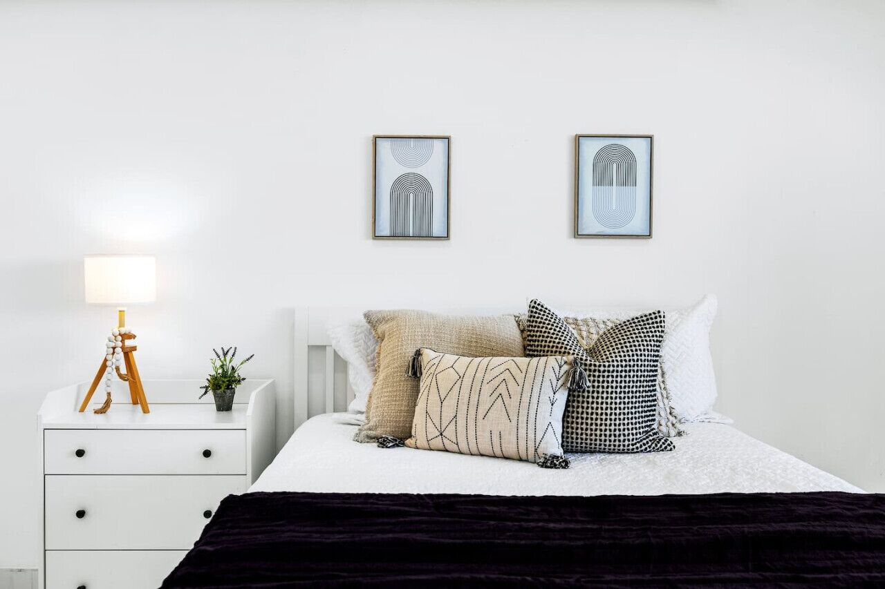 Una camera da letto ordinata con un letto, un comodino, una lampada e due opere d'arte appese alla parete.