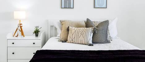 Una camera da letto ordinata con un letto, un comodino, una lampada e due opere d'arte appese alla parete.