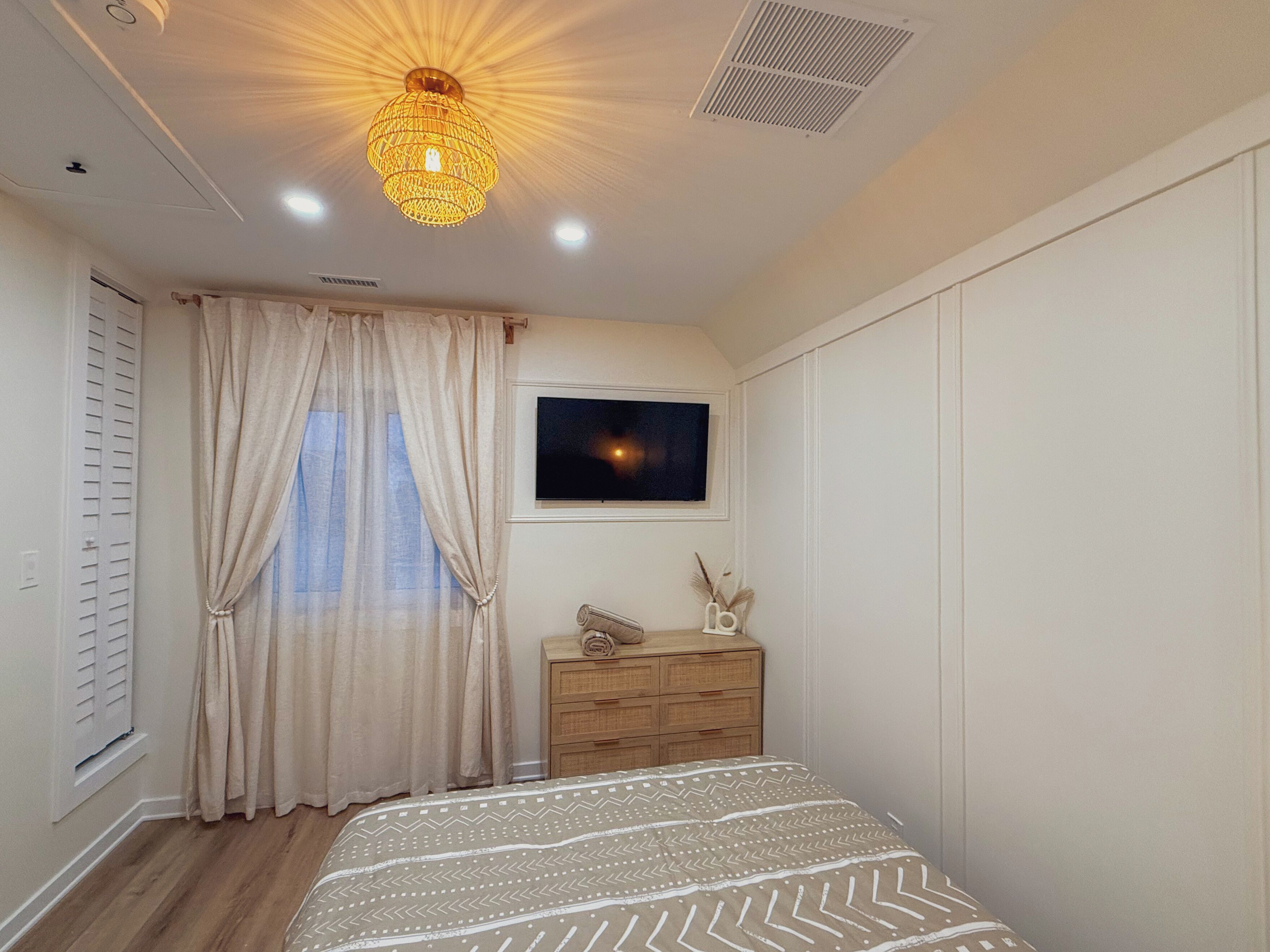Ein Schlafzimmer mit einem Bett, einem Schminktisch, einem Fenster mit Vorhängen, einer Deckenleuchte und einem an der Wand befestigten Fernseher.