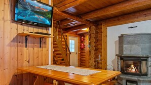 A wooden cabin interior with a wooden table, a fireplace, a television mounted on the wall, and a staircase leading to a door.