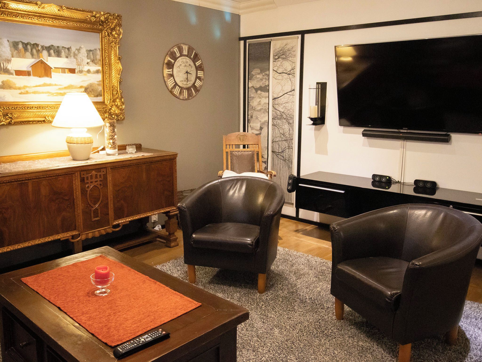 A living room with a wooden cabinet, two leather armchairs, a red rug, a remote control, a painting, a clock, and a flat-screen TV.