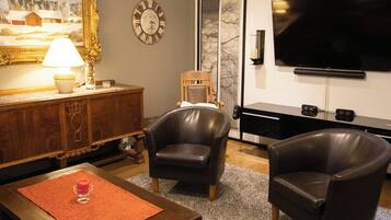 A living room with a wooden cabinet, two leather armchairs, a red rug, a remote control, a painting, a clock, and a flat-screen TV.