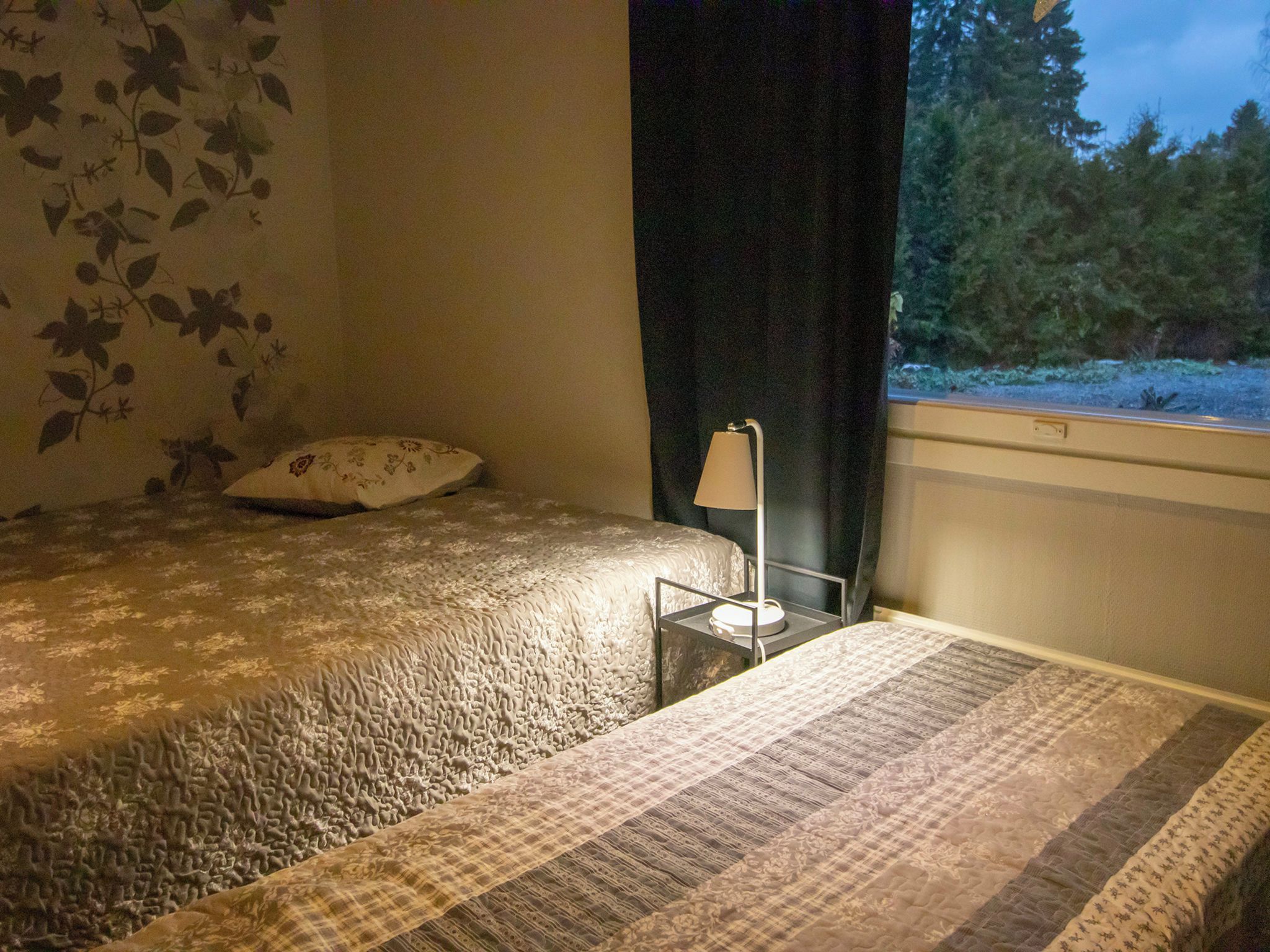 A bedroom with a bed, a bedside table with a lamp, and a window with a view of trees.