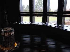 Huoneessa on ikkuna, josta avautuu näkymä vehreälle alueelle, takka, jossa on polttopuita, sekä metallinen astia, jossa on kiviä.