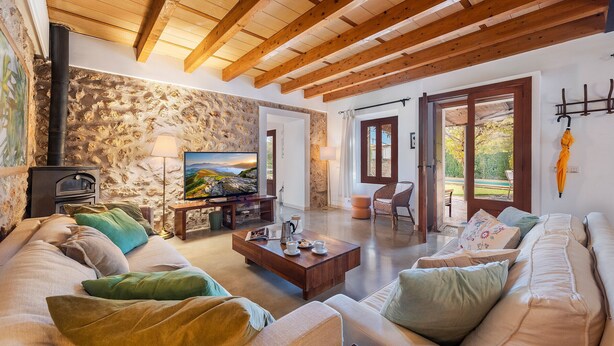 Ein gemütliches Wohnzimmer mit Steinwand, Holzbalken, Kamin, einer Couch, einem Couchtisch und einem Fernseher.