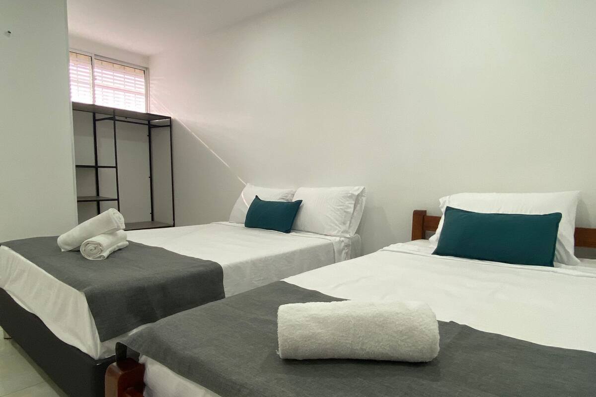 A hotel room with two single beds, each with a grey bedspread and white pillows, and a wooden headboard. There is a small shelf unit against the wall and a window with blinds.