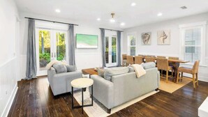 A modern living room with a sofa set, a dining table with chairs, and a view of greenery through the windows.