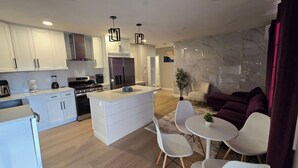 Eine moderne Küche mit weißen Schränken, einer zentralen Kochinsel und Edelstahlgeräten. Es gibt einen Essbereich mit weißen Stühlen und einem Tisch sowie eine Wohnbereich mit einer Couch und einer Pflanze.