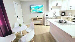 Eine moderne Küche mit weißen Schränken, einem eingebauten Geschirrspüler und einer Kaffeemaschine. Es gibt eine kleine Essecke mit weißen Stühlen und einem Tisch, sowie ein an der Wand befestigter Fernseher, der ein Landschaftsbild zeigt.