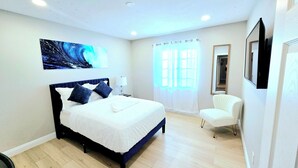 Ein Schlafzimmer mit einem Bett, einem Sessel, einem Gemälde und einem Fenster mit Vorhängen.