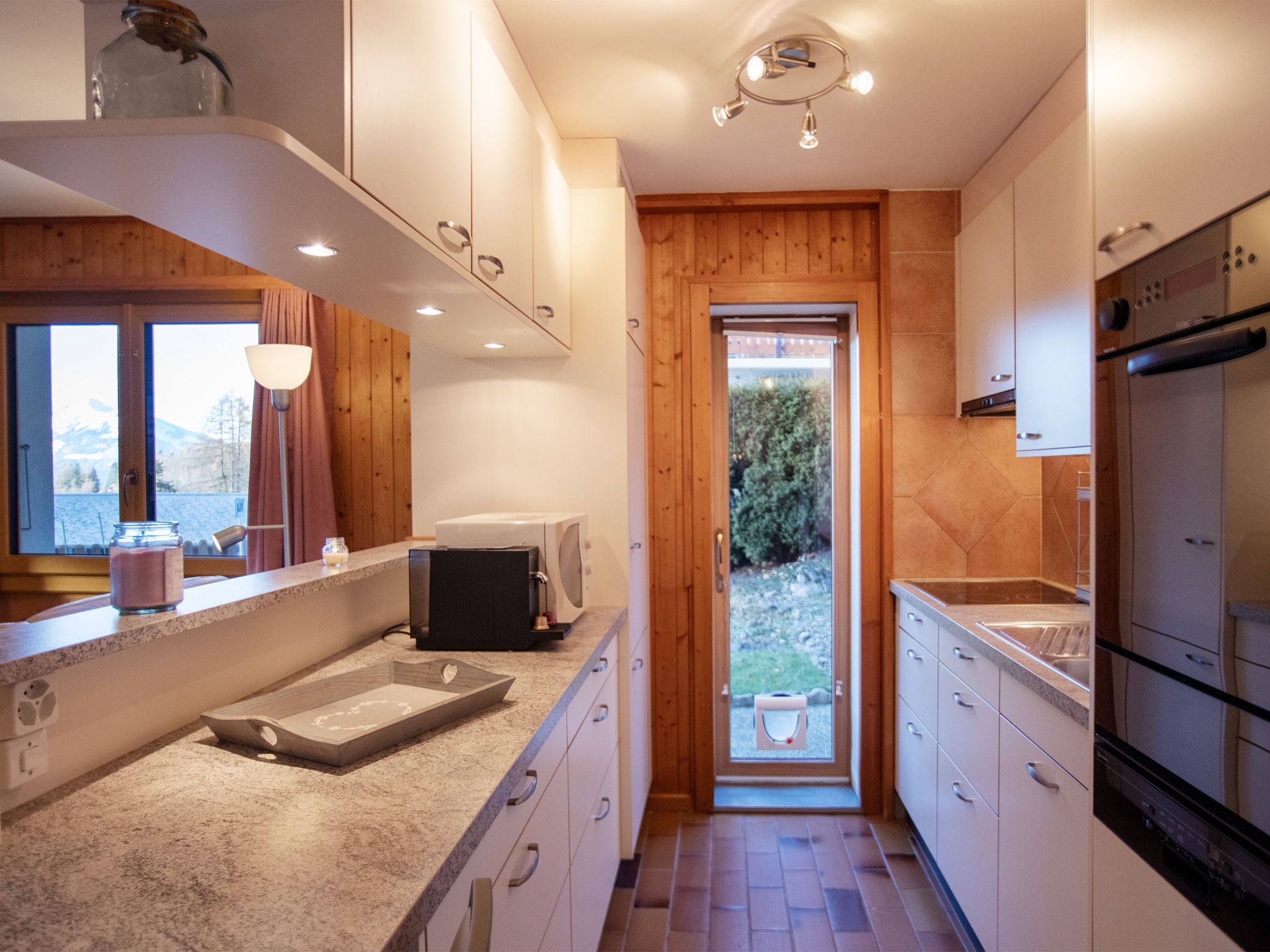 Une cuisine moderne avec une porte en bois donnant sur un balcon, des appareils électroménagers intégrés et une vue sur l’extérieur.