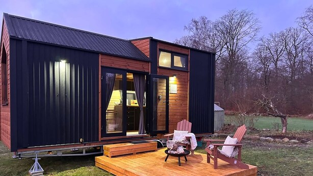 Ein modernes Haus mit einer Terrasse, zwei Adirondack-Stühlen und einer kleinen, durch die offene Tür einsehbaren Küche.