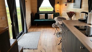 Ein gemütliches Blockhäuschen-Interieur mit Holzboden, einem grünen Sofa, einer kleinen Küchenzeile und Blick ins Freie.