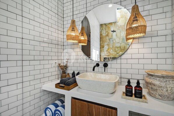 Une salle de bain aux murs recouverts de carreaux blancs, un grand miroir rond, un lavabo blanc avec deux flacons de savon liquide et un grand bac en pierre.