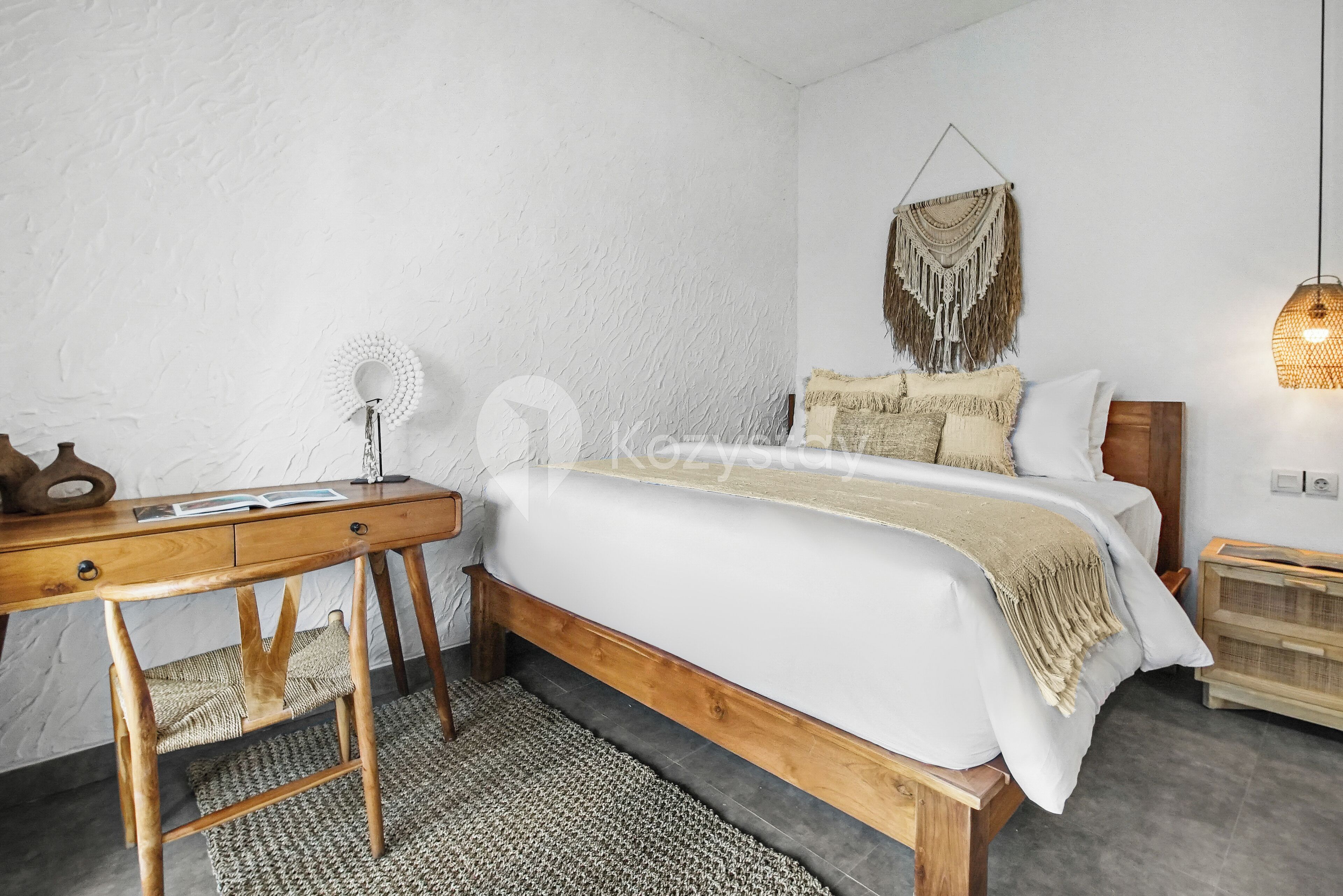 A bedroom with a wooden bed, a desk with a chair, a macrame wall hanging, and a wall-mounted lamp.
