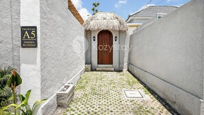 A narrow pathway leading to a thatched-roof entrance with a wooden door, surrounded by white walls and a tiled floor. - Kalea Villa by Kozystay - Ungasan (Ungasan)