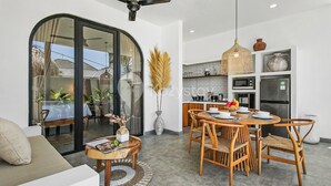 A modern living room with a sofa, a dining table with chairs, a kitchen area with appliances, and a large window with a view of the outside. - Kalea Villa by Kozystay - Ungasan (Ungasan)
