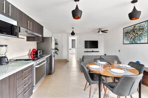 A modern kitchen with stainless steel appliances, a dining area with a wooden table and chairs, and a living area with a sofa and a TV.