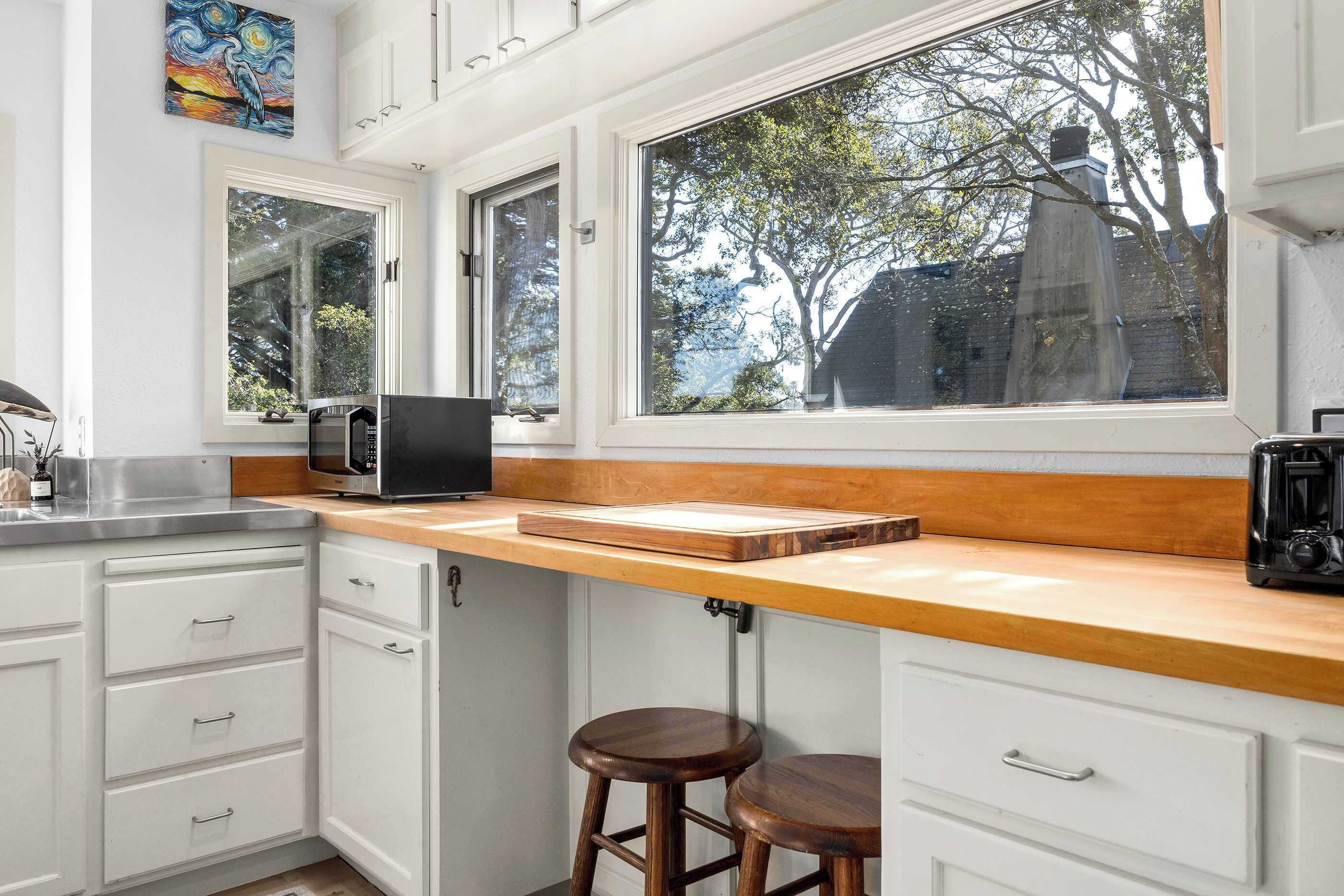 A kitchen with white cabinets, a wooden countertop, a microwave, a toaster, and a window with a view of trees and a building.