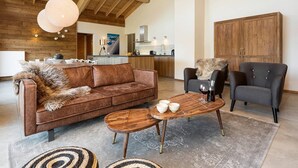 Amplia sala de estar con un sofá de cuero marrón, una mesa de centro de madera y un sillón gris. Se ve una cocina al fondo.