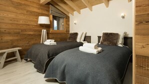 Una habitación acogedora con paredes de madera y techo inclinado, con dos camas vestidas con ropa de cama oscura y almohadas mullidas, una mesita de noche pequeña y una lámpara.