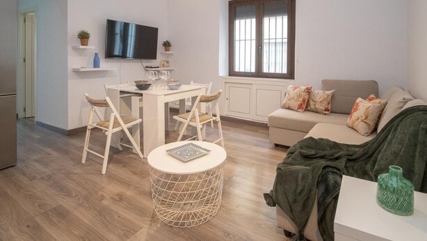 A modern living room with a sofa, a dining table with chairs, a TV mounted on the wall, and a window with blinds.