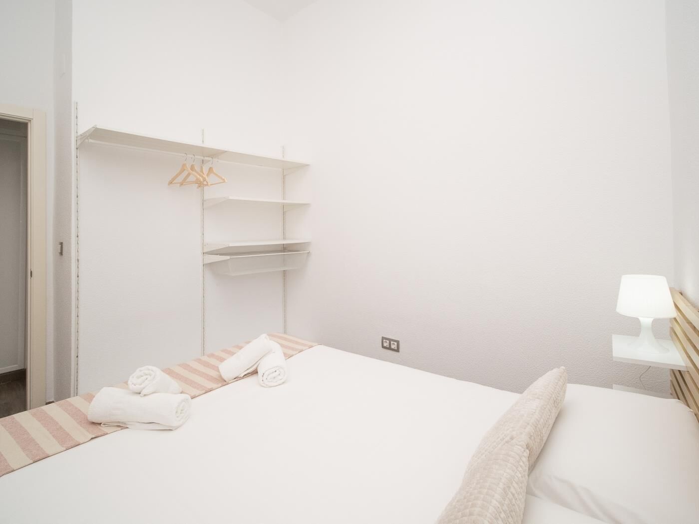 A minimalist hotel room with a bed, a bedside table with a lamp, and a wardrobe with hanging clothes.