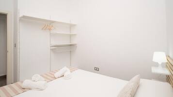 A minimalist hotel room with a bed, a bedside table with a lamp, and a wardrobe with hanging clothes.
