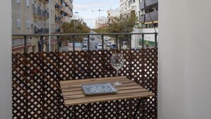 A balcony with a wooden table and a wine glass, overlooking a street with buildings and parked cars.