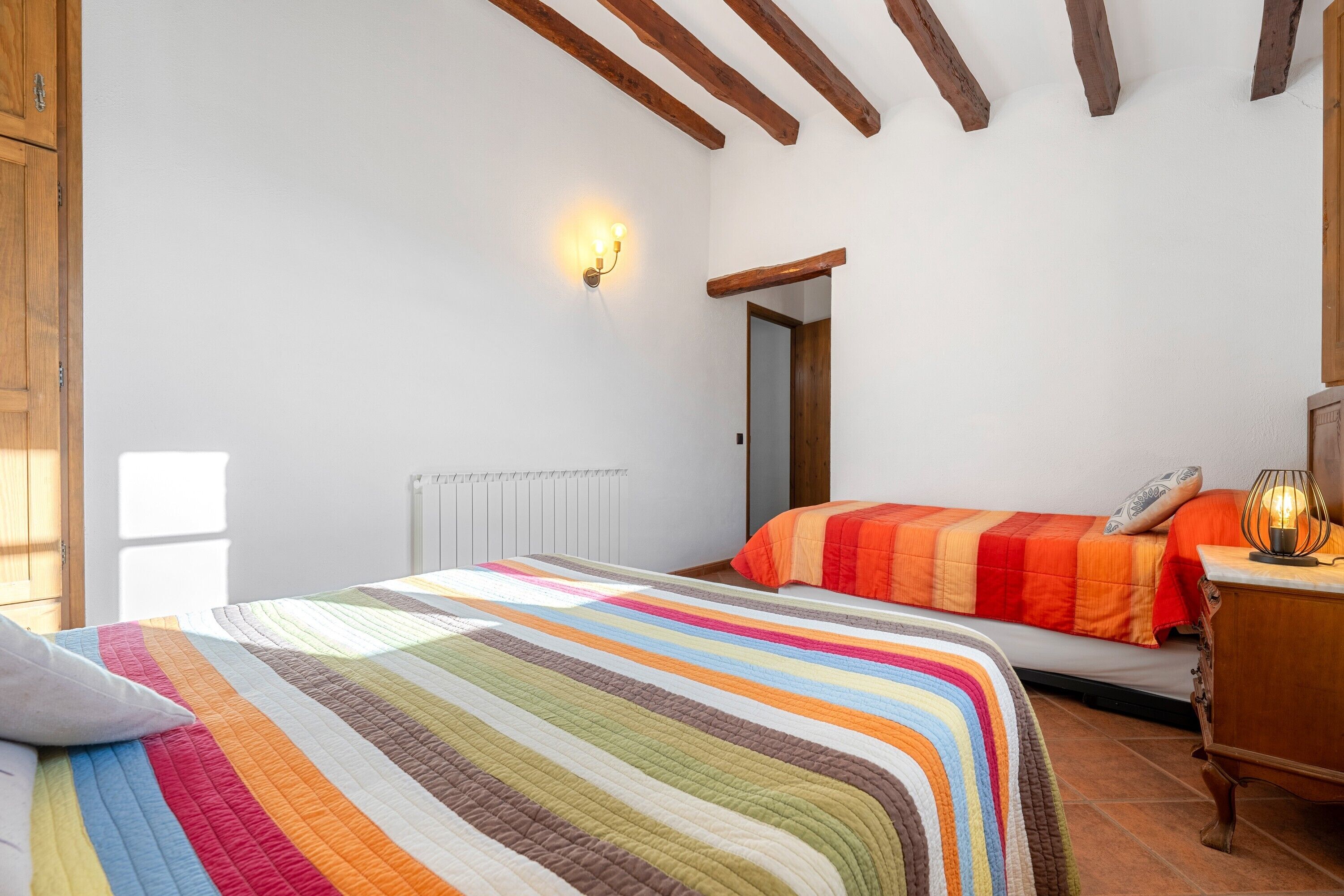 A bedroom with a striped bedspread, a wooden dresser, a nightstand with a lamp, and a radiator.