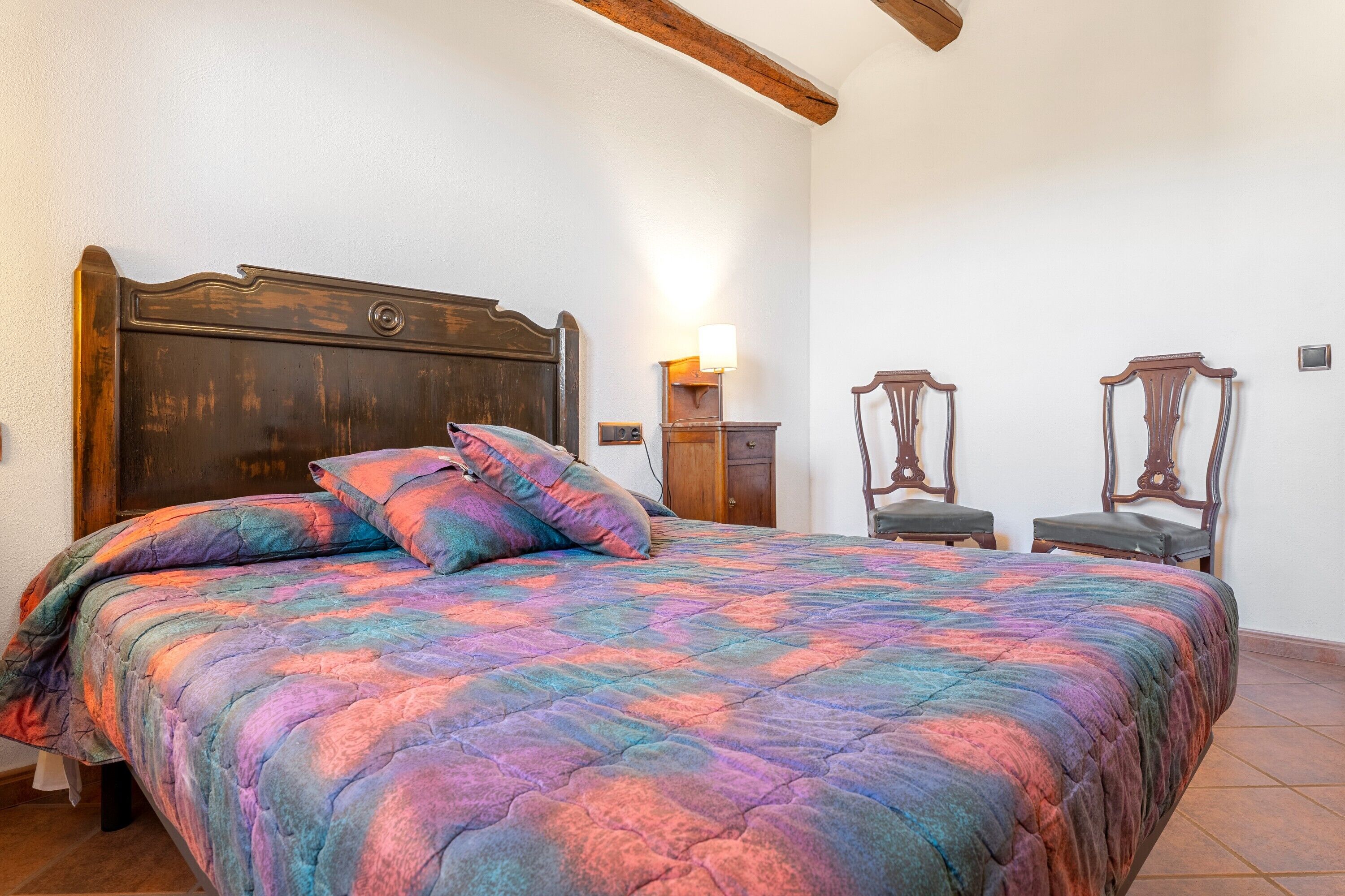 A bedroom with a bed, two chairs, a nightstand, and a wall-mounted lamp.