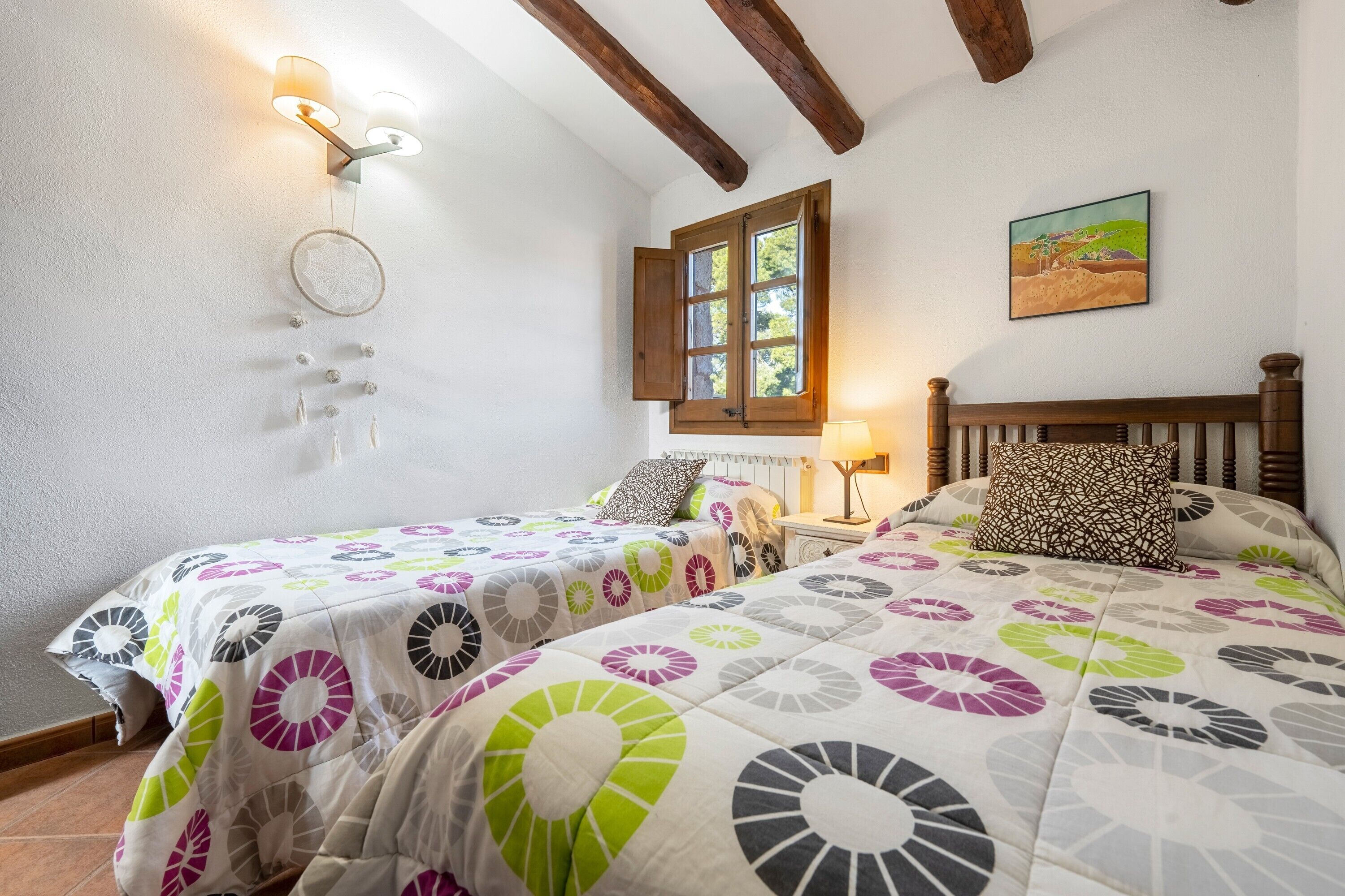 A bedroom with two beds, a window, a wall-mounted light, and a painting.
