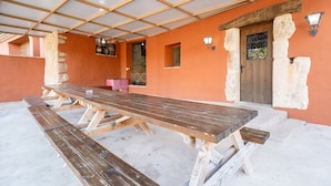 Un espace extérieur abrité, aménagé pour les repas, avec une longue table en bois et des bancs, un mur en pierre et une porte surmontée d’un portail en métal.
