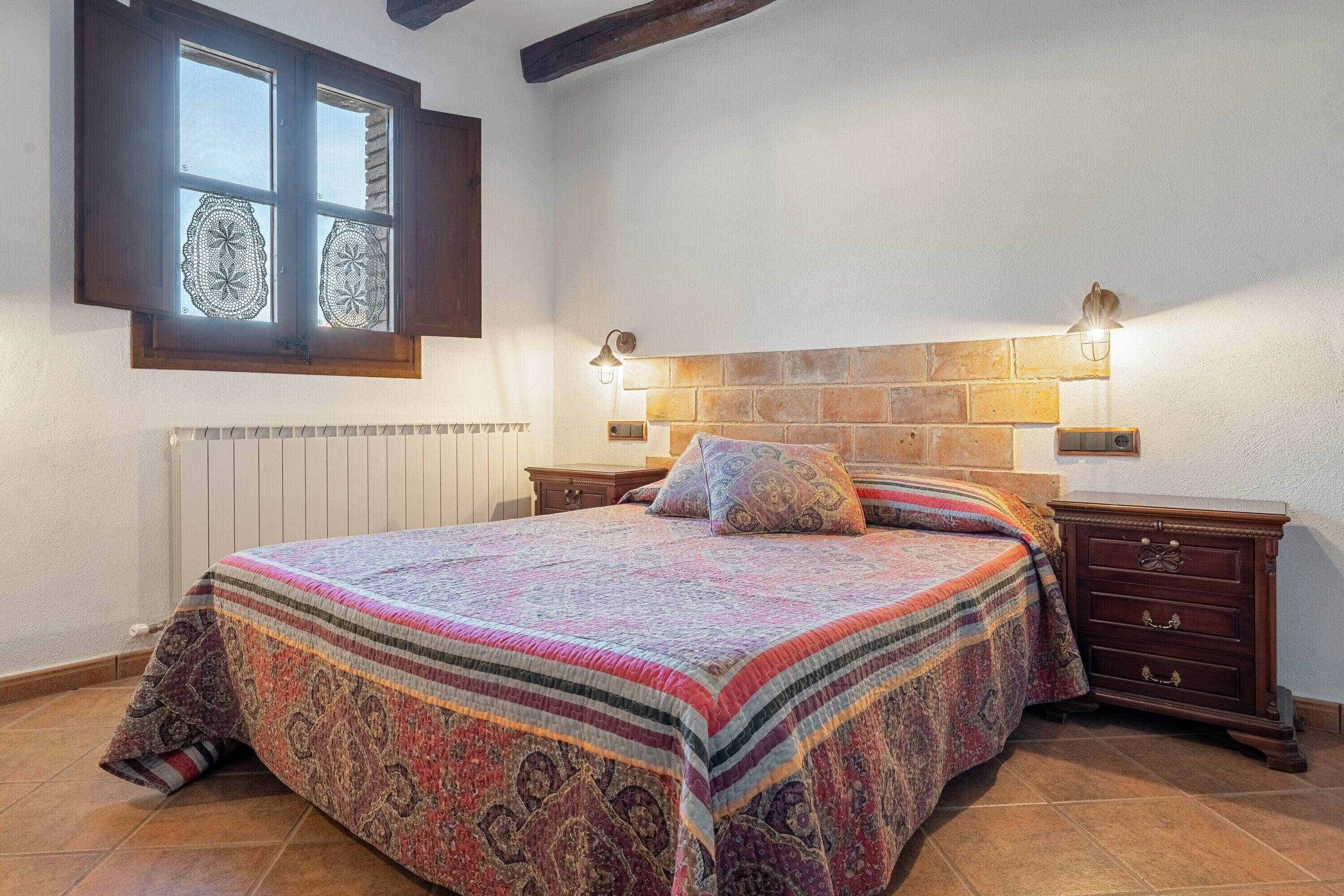 Een slaapkamer met een groot bed, een raam met kantgordijnen, een hoofdbord met baksteenmotief, en twee wandlampen.