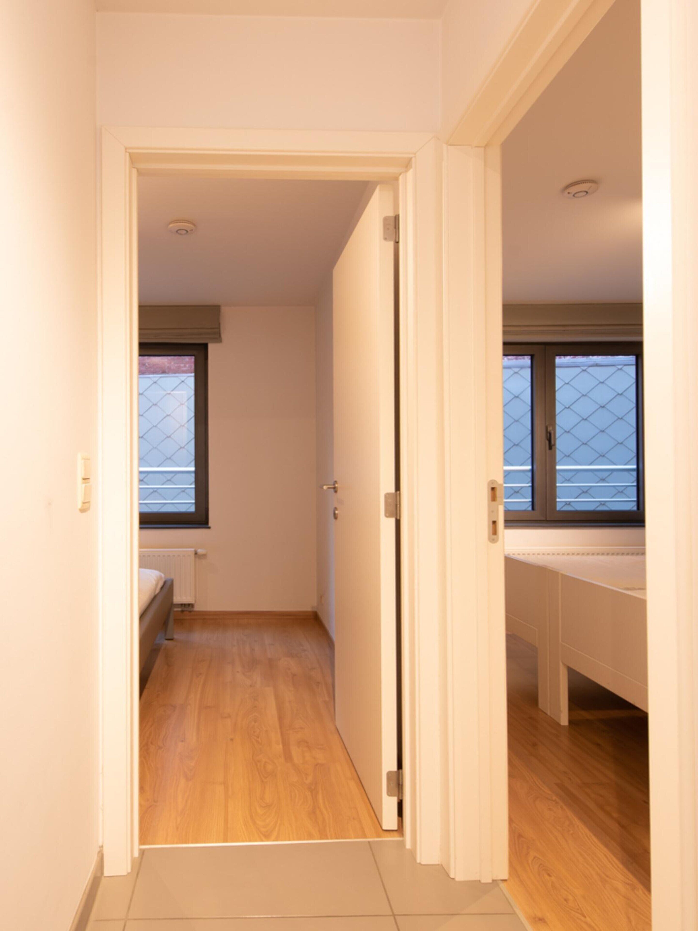 A narrow hallway with wooden flooring, a bed on the left, and a window with blinds on the right.