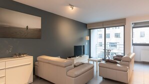 A modern living room with a large window, a flat-screen TV, and a minimalist design. - Apartment in Ostend Near the Seafront (Ostend)