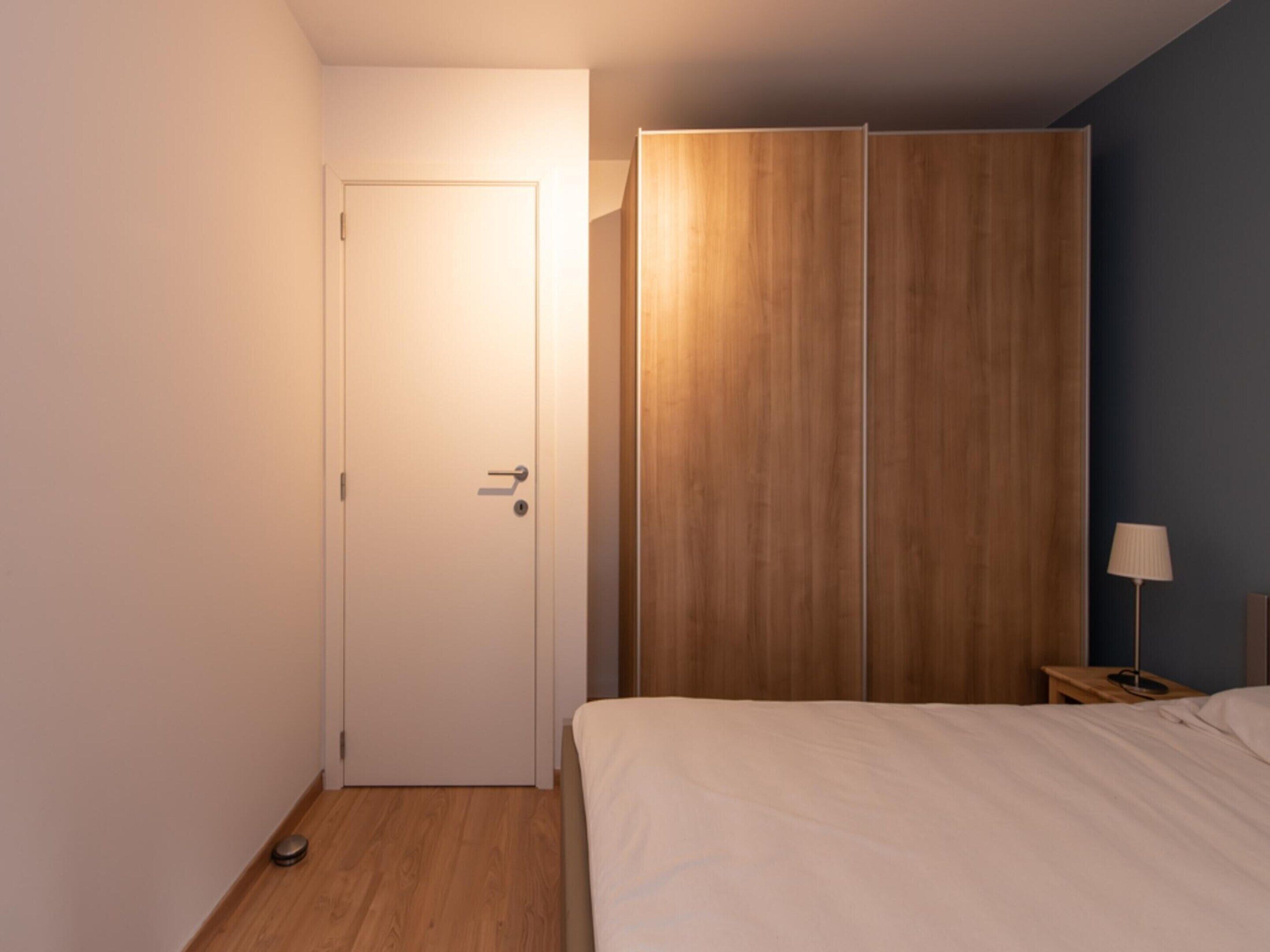 Un dormitorio con cama, armario de madera, mesita de noche con lámpara y una puerta.