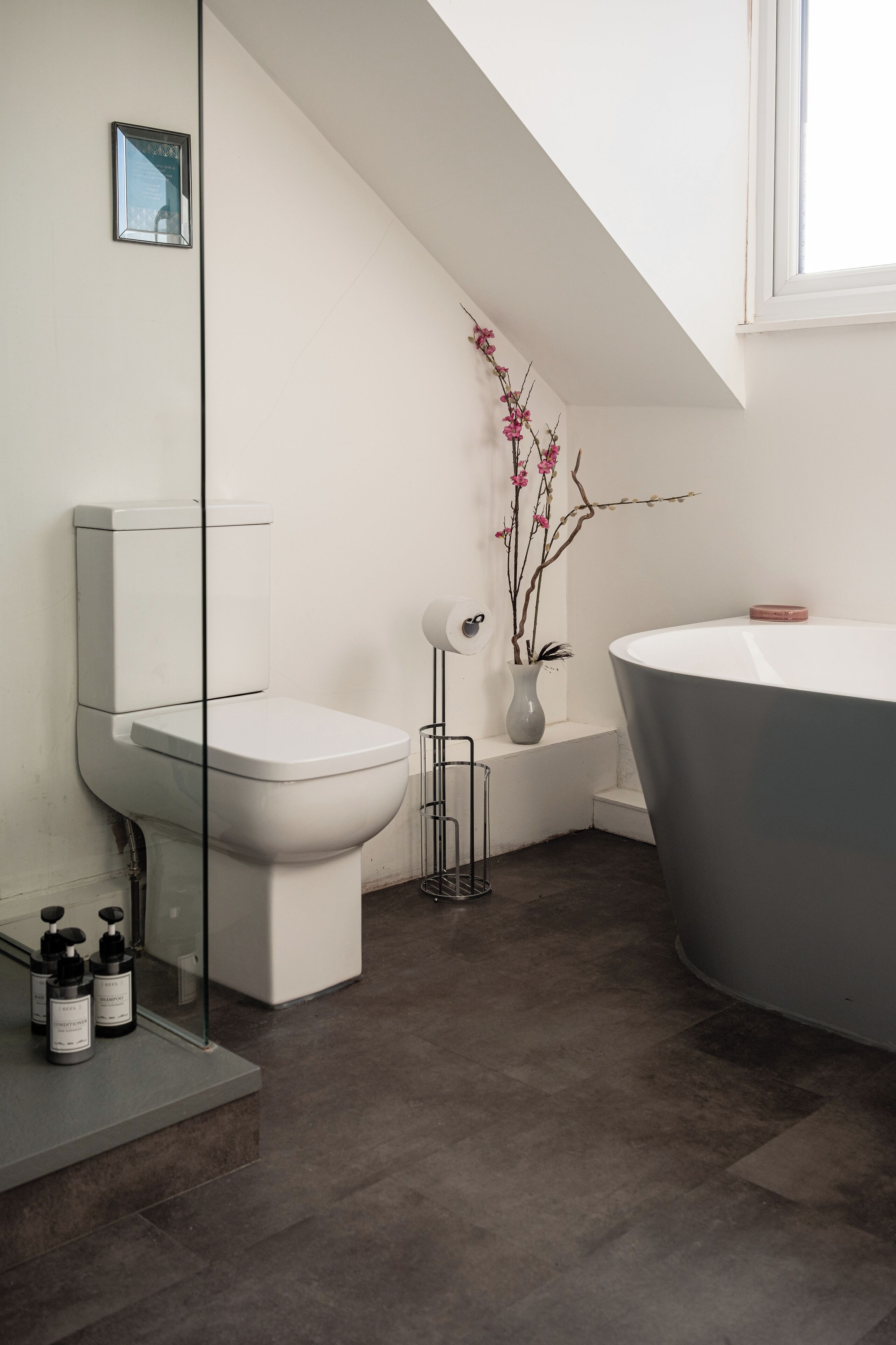 A modern bathroom with a toilet, bidet, and bathtub. There is a vase with flowers on the wall and a small window above the bathtub.