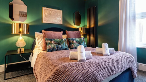 Una cama bien hecha con un edredón de punto, dos lámparas de noche, una obra de arte enmarcada y una ventana con cortinas.