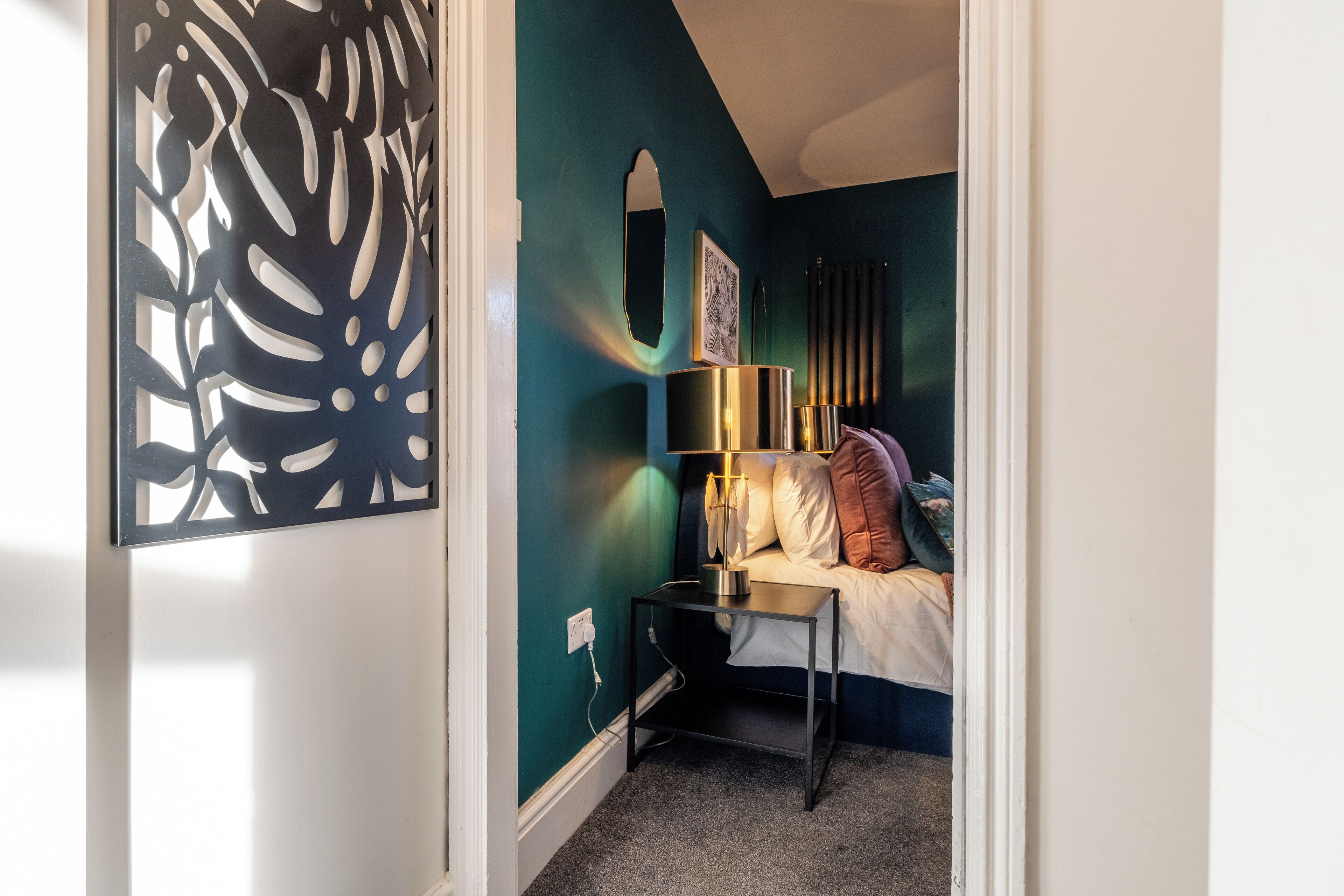 A bedroom with teal walls, a bed with white bedding, a bedside table with a lamp, and a decorative wall piece.