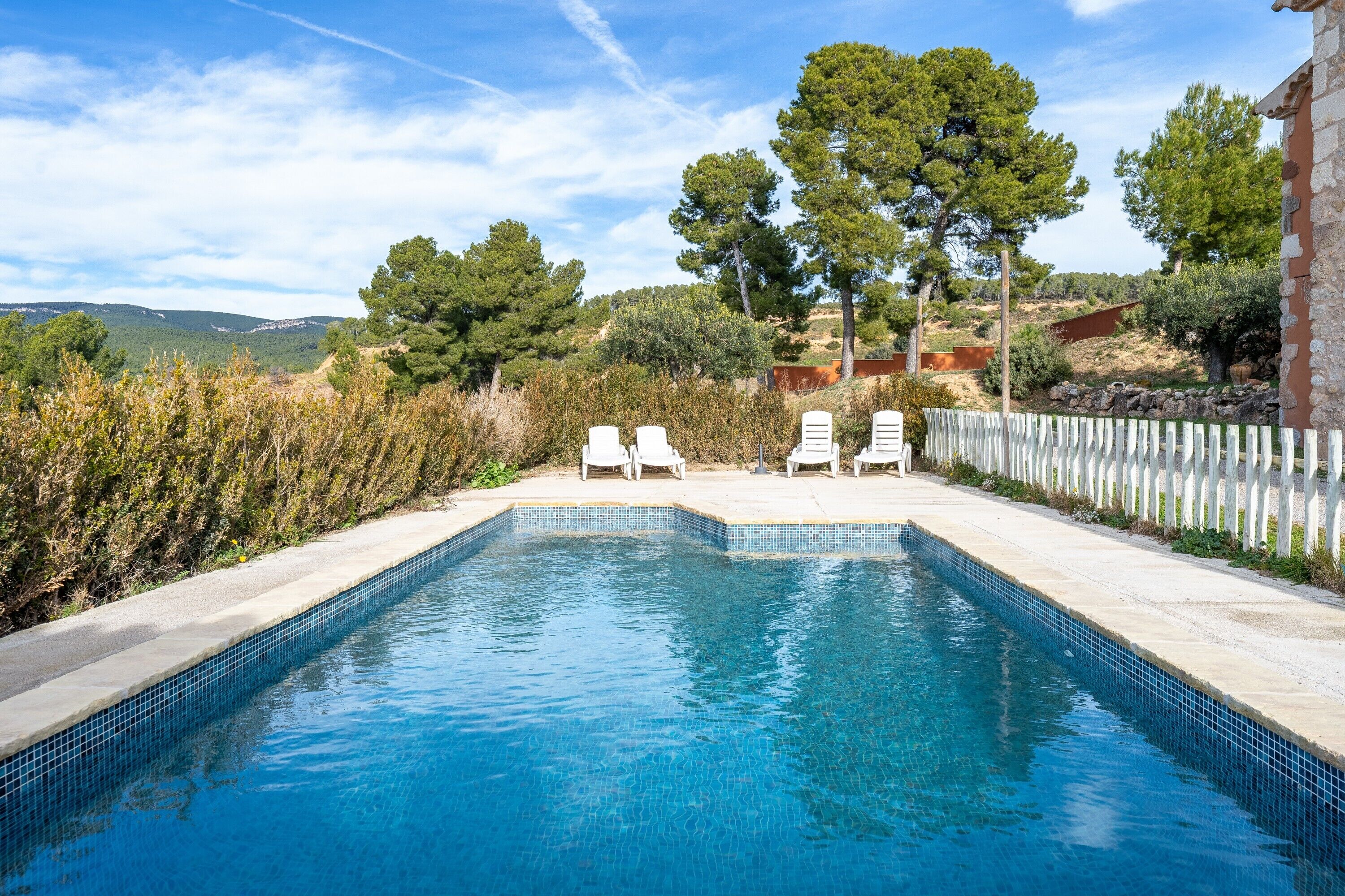 Una piscina rectangular con agua azul cristalina, rodeada por una cerca blanca y dos sillas reclinables blancas.