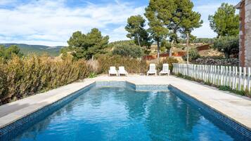 Una piscina rectangular con agua azul cristalina, rodeada por una cerca blanca y dos sillas reclinables blancas.